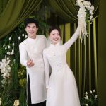 Cặp đôi mặc áo dài cưới trắng tinh khôi tạo dáng duyên dáng trước phông nền xanh sang trọng.