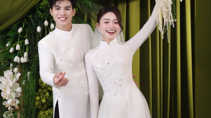 Cặp đôi mặc áo dài cưới trắng tinh khôi tạo dáng duyên dáng trước phông nền xanh sang trọng.