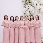 Nhóm các cô gái mặc áo dài hồng duyên dáng đứng trước phông nền hoa trắng.